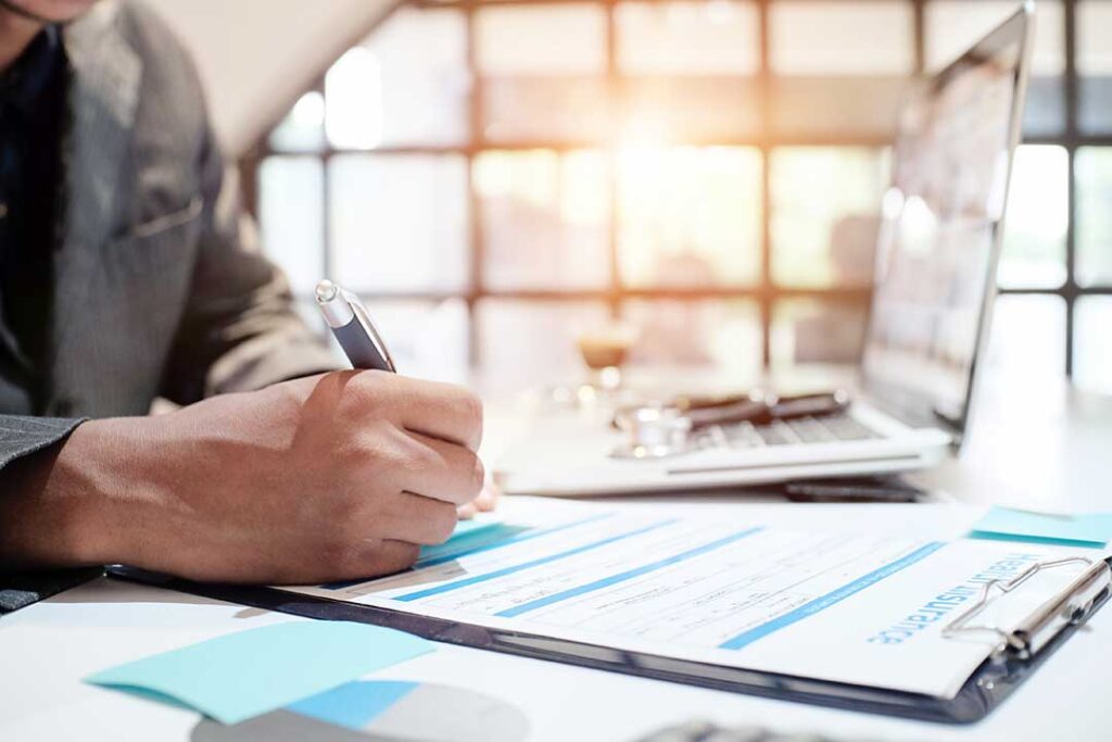 A person reading through paperwork at a home office desk with policy documents and a pen in hand.